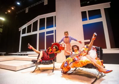 Play scene with two women in colorful dresses lounging in chairs, another in an apron standing behind them. From the opera "La cenerentola" with the Manitoba Opera 2022.