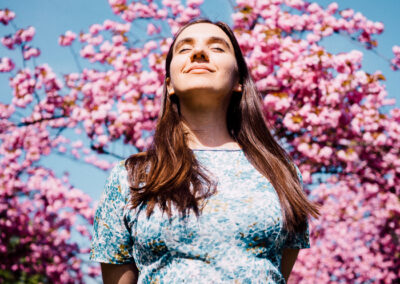A woman in a blue floral dress stands outdoors, smiling and looking up. Behind her are blooming pink cherry blossom trees under a clear blue sky, creating a vibrant spring atmosphere.
