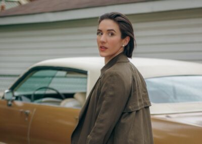 Woman in olive trench coat and black boots in front of vintage yellow car.