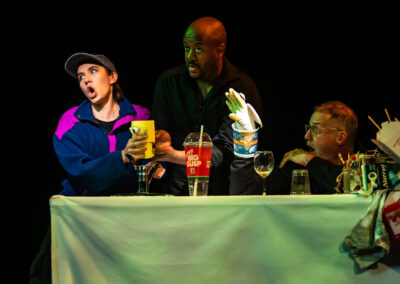 Three actors performing on stage with props at a table.