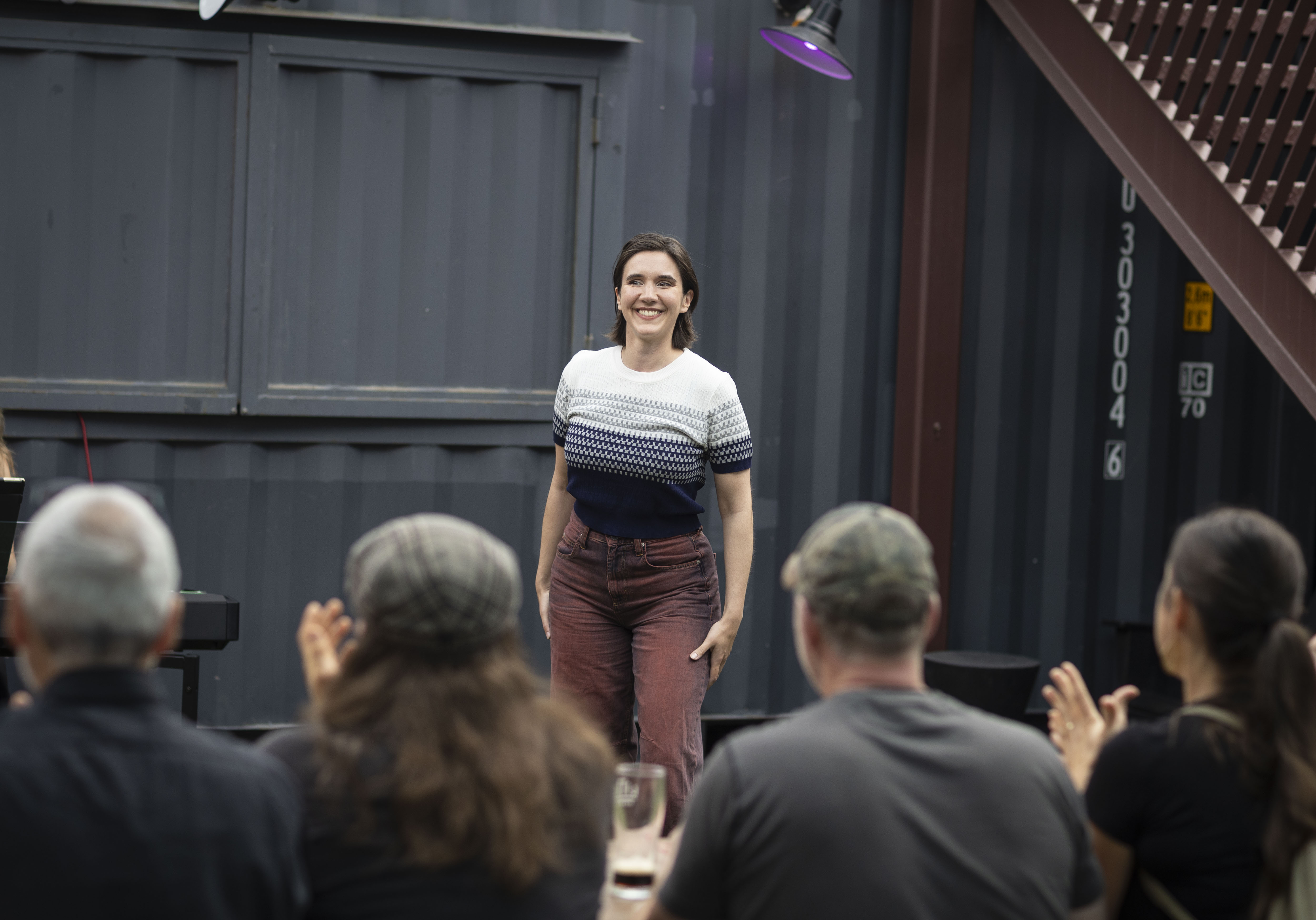 Woman in a patterned sweater and rust-colored pants smiles at an audience.